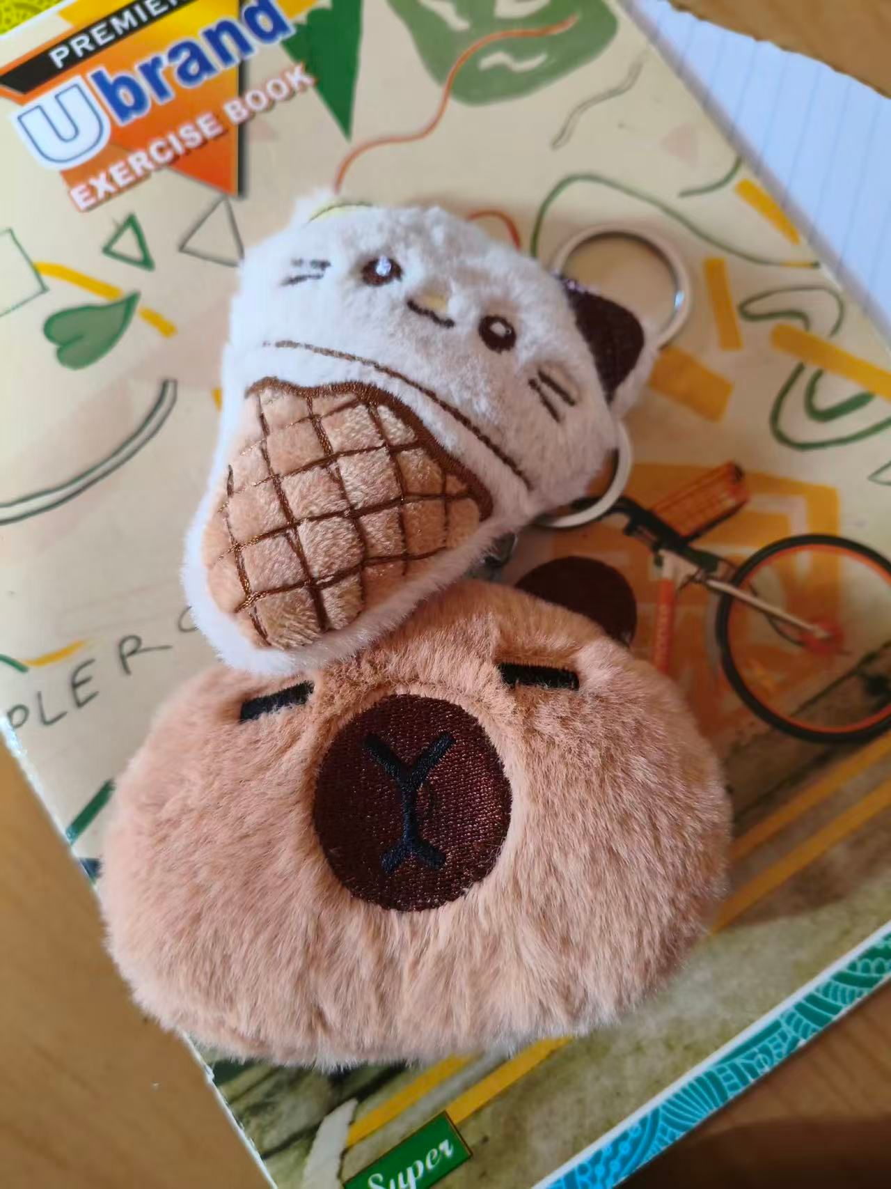 Two plush animal keychains — a cat and a capybara — resting on the cover of a school exercise book.