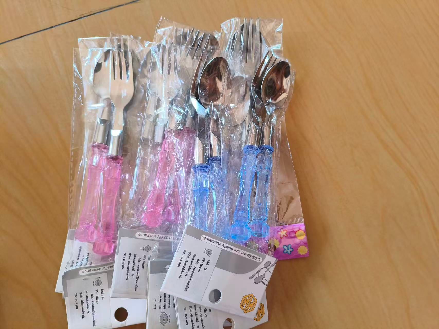 Packaged stainless-steel fork-and-spoon sets with translucent pink and blue handles, laid out on a table.