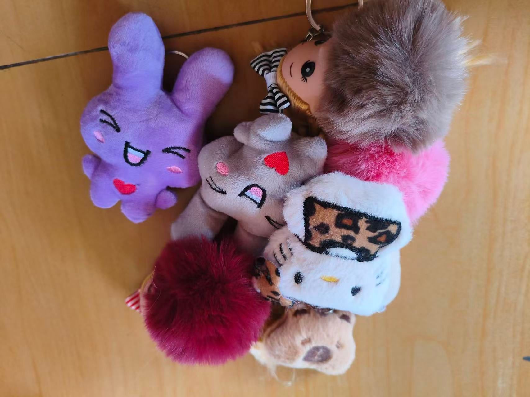 A cluster of plush keychains and pom-pom charms in several colours and characters, gathered on a wooden floor.