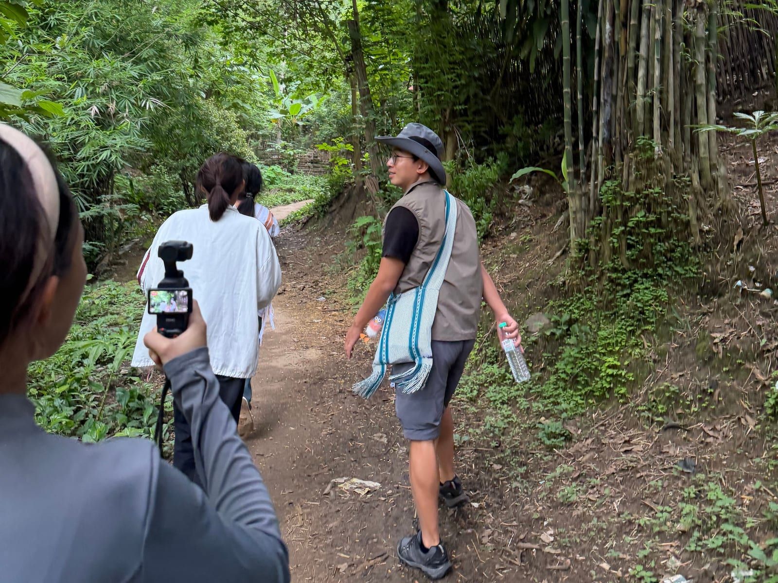 Three people walk along a dirt path under tall bamboo and trees. A young man in a wide-brimmed hat with a woven shoulder bag glances to the side, and someone in the foreground holds up a small handheld camera.