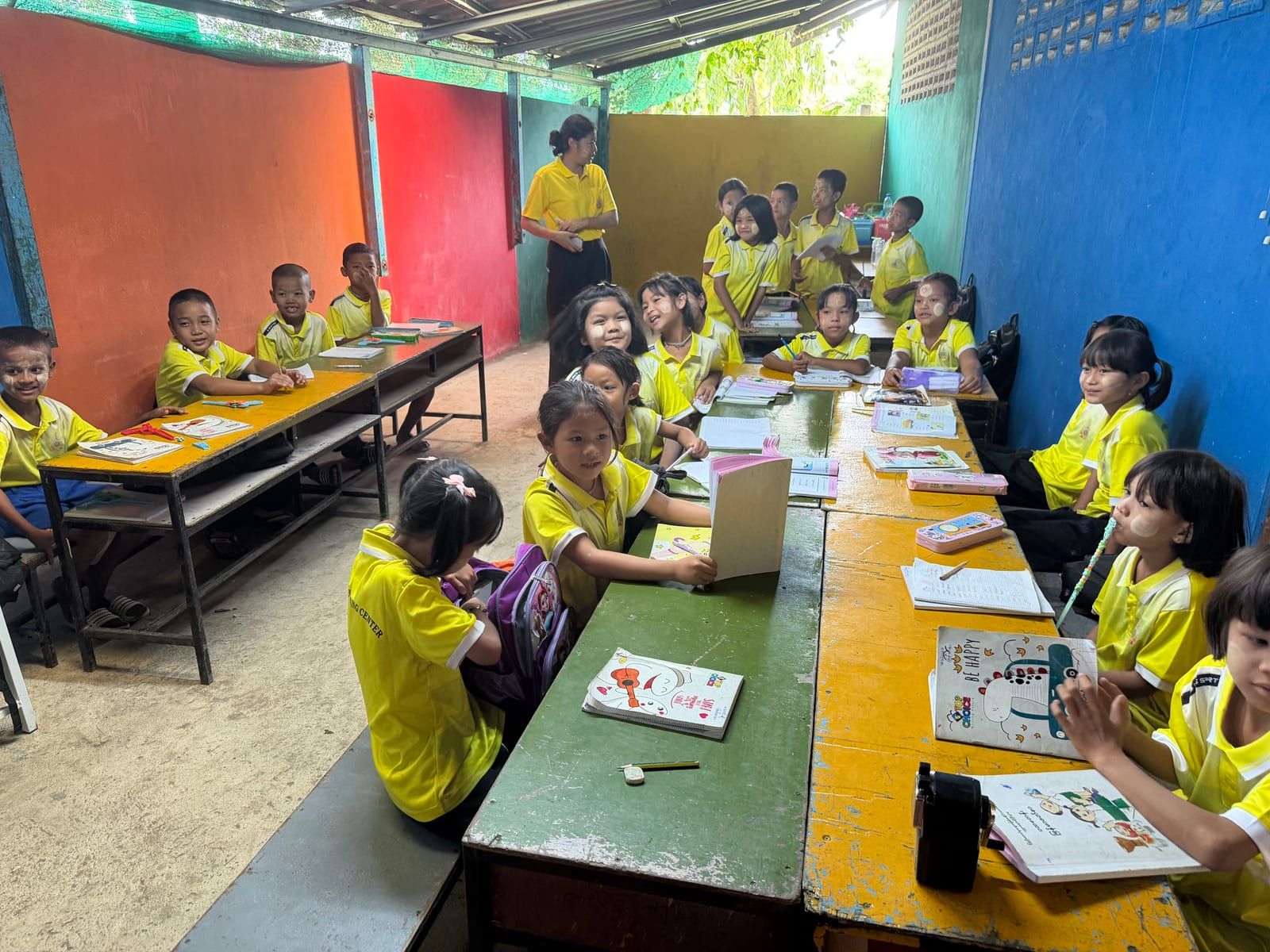 About twenty children in yellow polo shirts sit or stand at long green and yellow tables with notebooks, many smiling at the camera. A woman in a yellow shirt stands at the back of a room with orange, red, yellow, green and blue walls.
