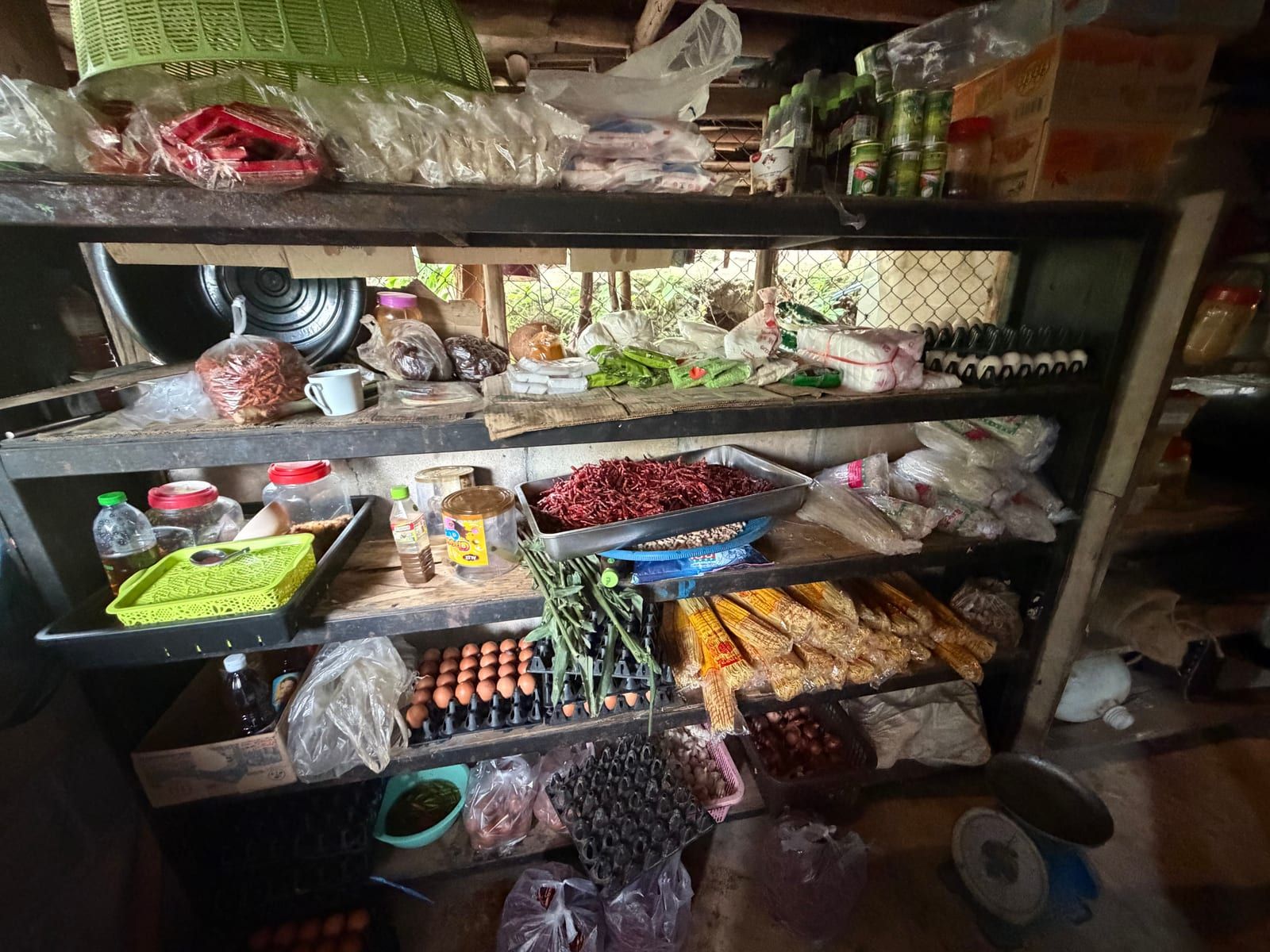 Wooden shelves stacked with trays of eggs, a tray of dried red chillies, bundles of yellow noodles, jars, bottled sauces and bagged dry goods, with a weighing scale on the floor.